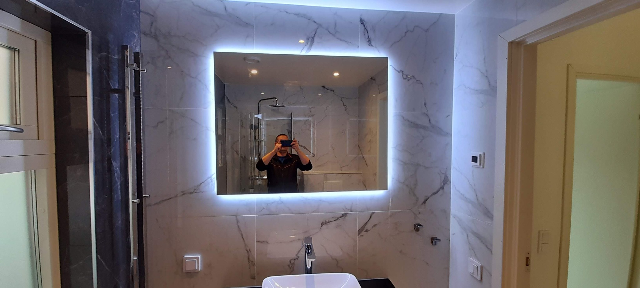 Nowoczesna łazienka z marmurowymi płytkami i podświetlanym lustrem, w odbiciu widoczna osoba robiąca zdjęcie telefonem, bateria umywalkowa w minimalistycznym stylu.