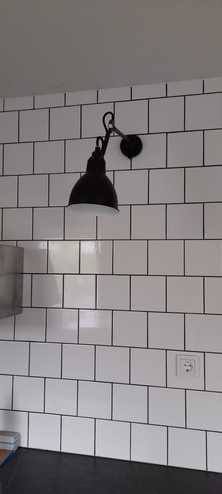 Czarna, metalowa lampa ścienna w stylu industrialnym zamontowana na białej ścianie z płytek typu metro z czarną fugą, widoczne gniazdko elektryczne i fragment blatu.