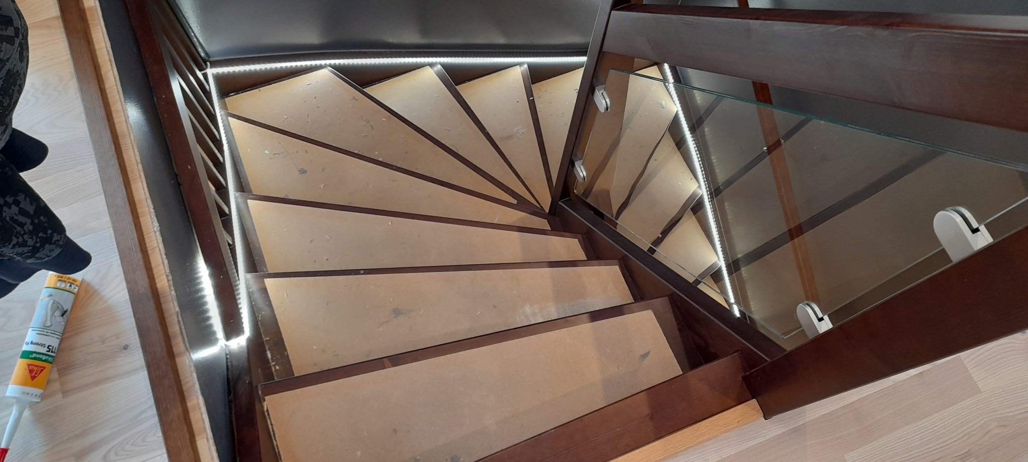 Klatka schodowa spiralna z drewnianymi stopniami i szklaną balustradą, podświetlona taśmą LED, widok z góry w trakcie montażu, obok tuba z klejem montażowym.