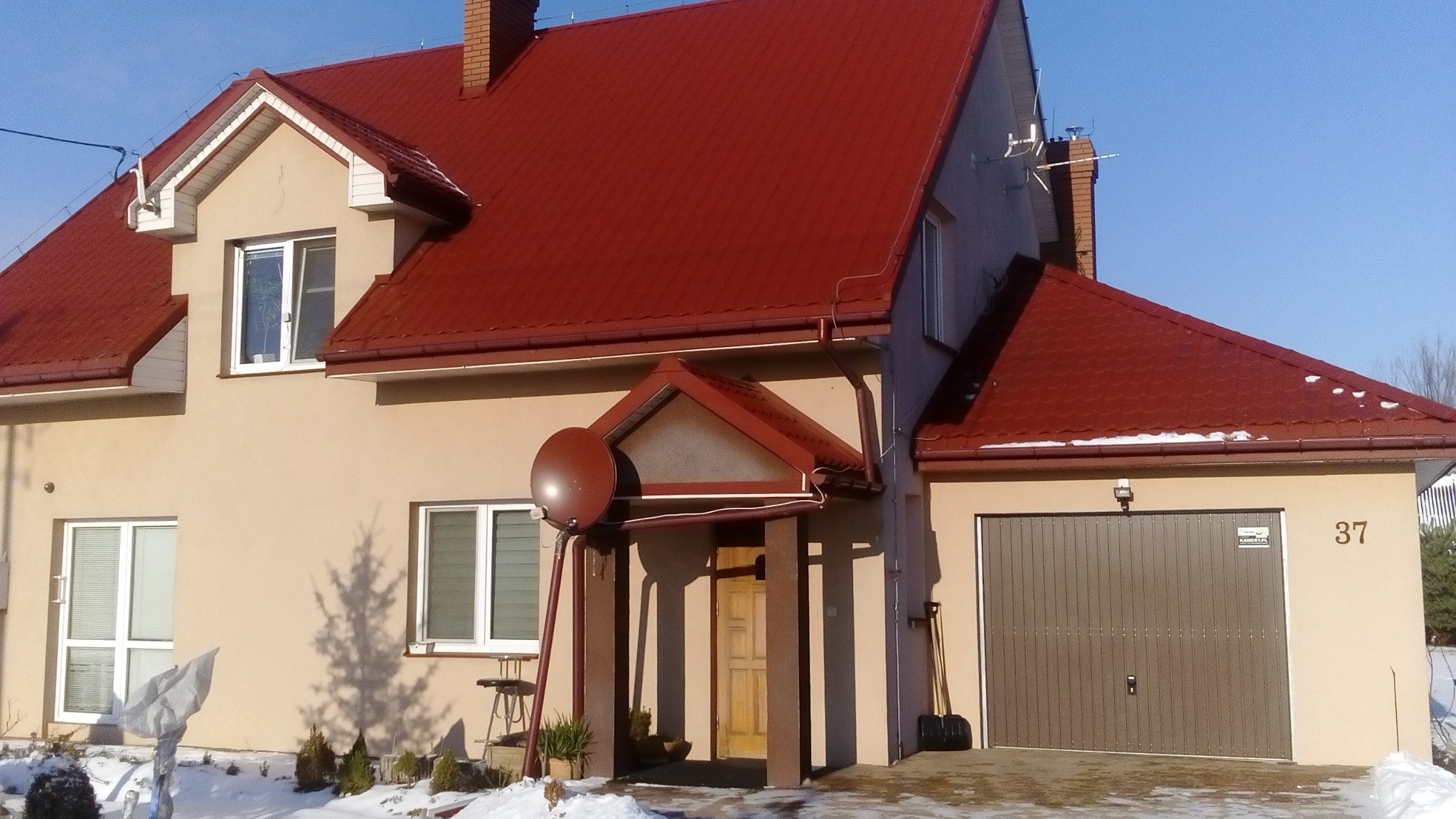 Dom jednorodzinny z czerwoną dachówką, antena satelitarna zamontowana przy wejściu, zimowy krajobraz z odśnieżonym podjazdem.