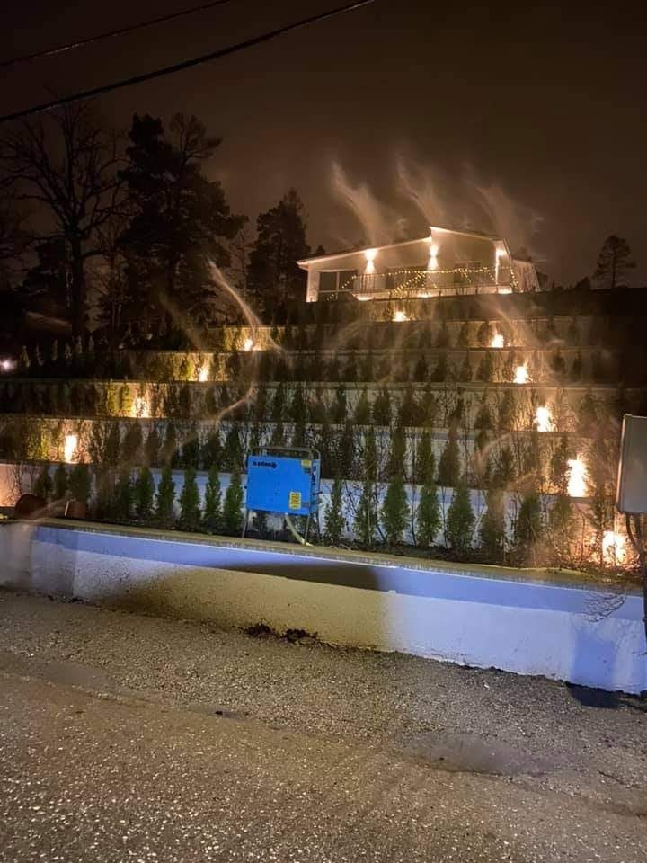 Nocne ujęcie domu z podświetlonymi schodkowymi rabatami, system nawadniania w akcji, niebieski agregat pompowy widoczny na pierwszym planie.