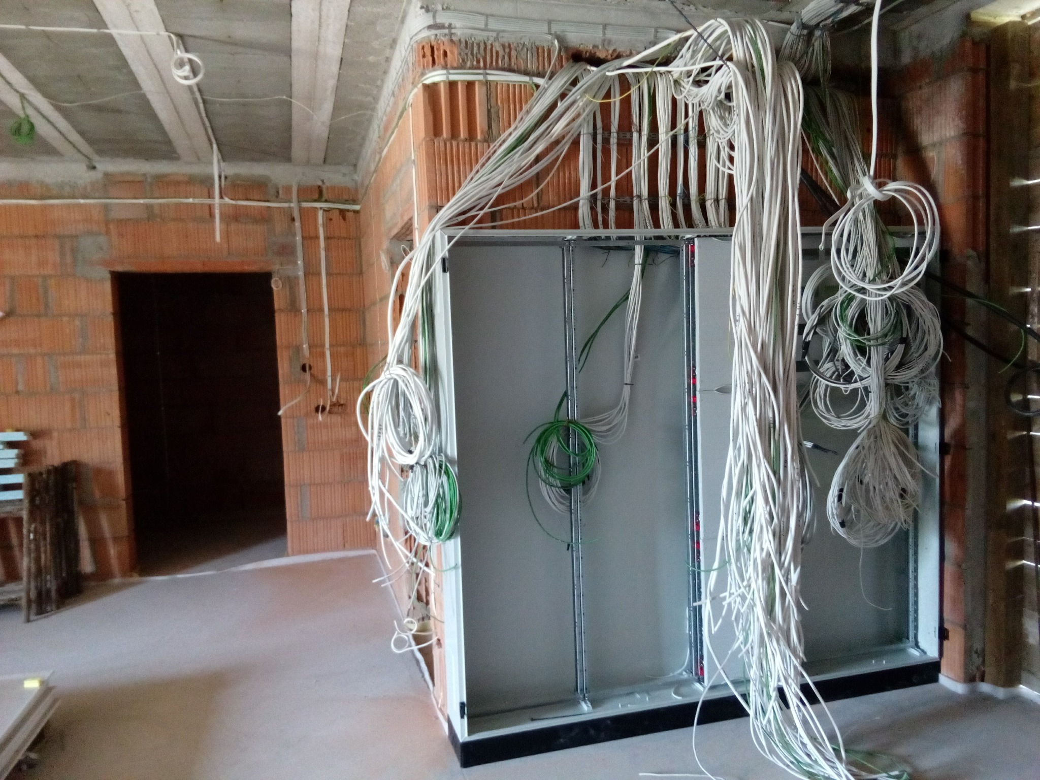 Rozdzielnica elektryczna w surowym stanie z licznymi przewodami wychodzącymi z góry i boków, widoczne ściany z czerwonej cegły i surowy strop.