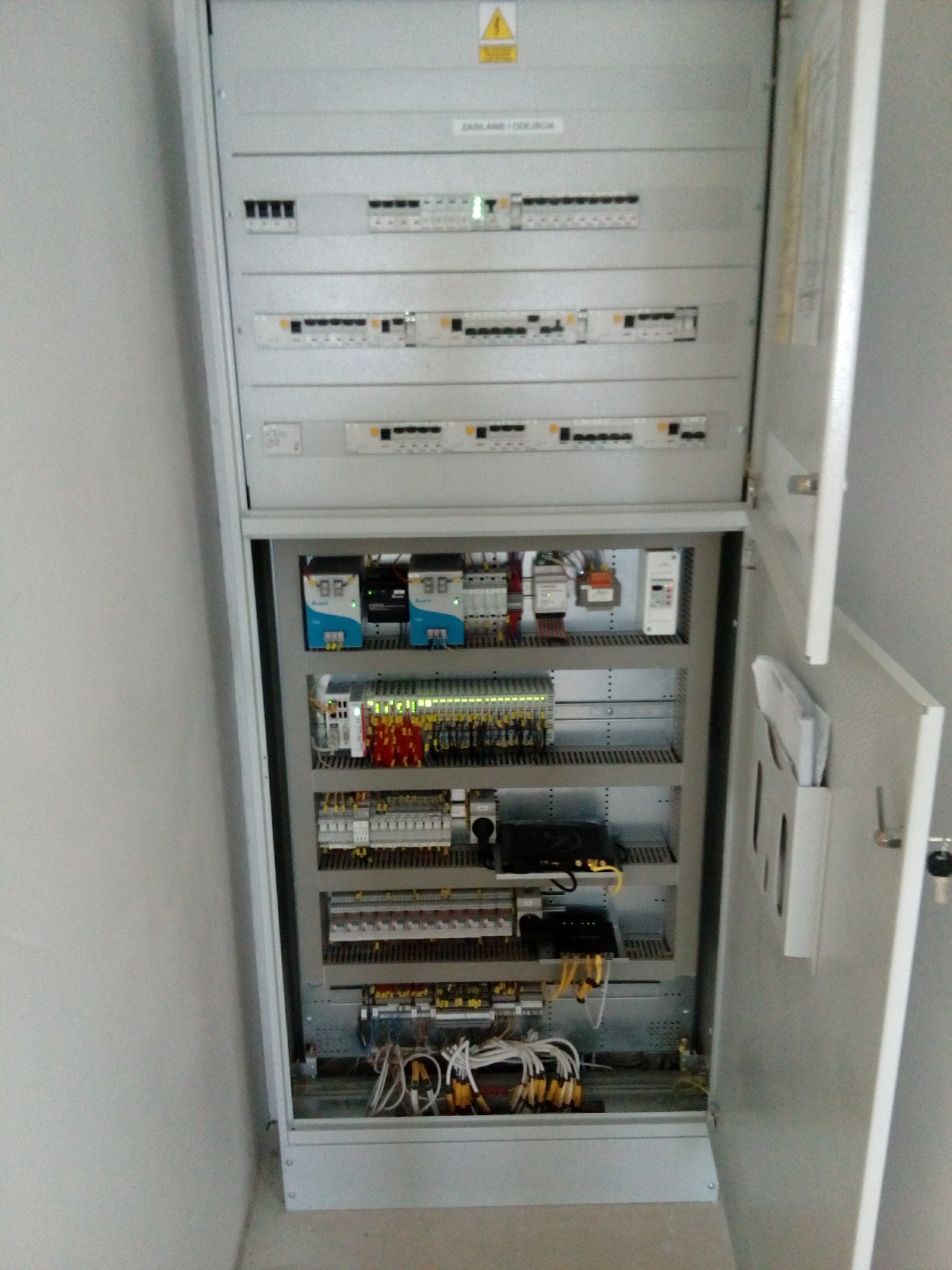 Otwarta szafa elektryczna z widocznymi bezpiecznikami, sterownikami PLC, przewodami i listwami zaciskowymi, umieszczona na szarej ścianie.