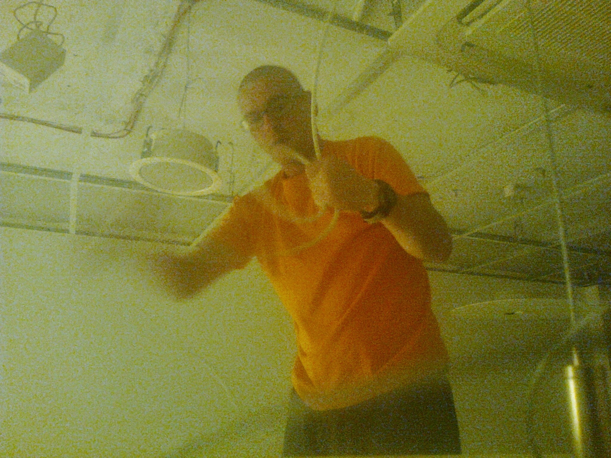 Elektryk w pomarańczowej koszulce, podczas montażu oświetlenia w pomieszczeniu z widoczną instalacją elektryczną i lampami sufitowymi.