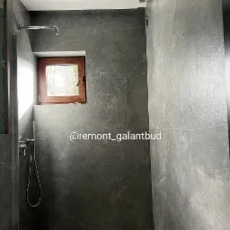 Szara łazienka z oknem w drewnianej ramie, wykończona płytami imitującymi kamień, widoczna armatura prysznicowa i fragment toalety.