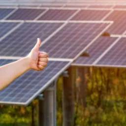 Ręka z uniesionym kciukiem na tle rzędu paneli fotowoltaicznych zamontowanych na konstrukcji, w tle zieleń i promienie słońca.