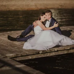 Para młoda siedzi na drewnianym pomoście nad wodą, w tle widoczne drzewa, panna młoda w białej sukni, pan młody w granatowym garniturze z muszką.