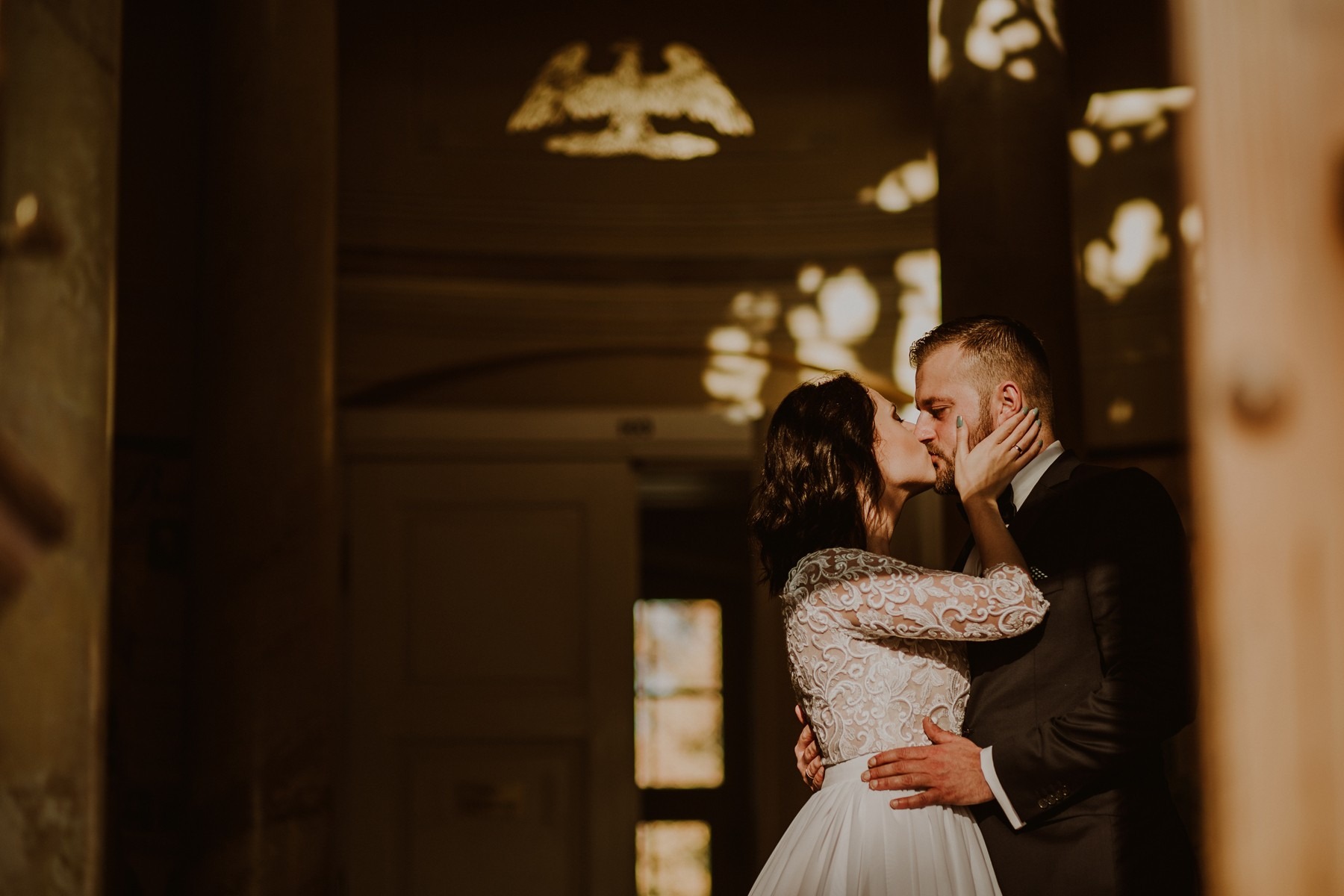 Pocałunek młodej pary w korytarzu z dekoracyjnymi cieniami na ścianach, panna młoda w koronkowej sukni z długim rękawem, pan młody w czarnym garniturze.