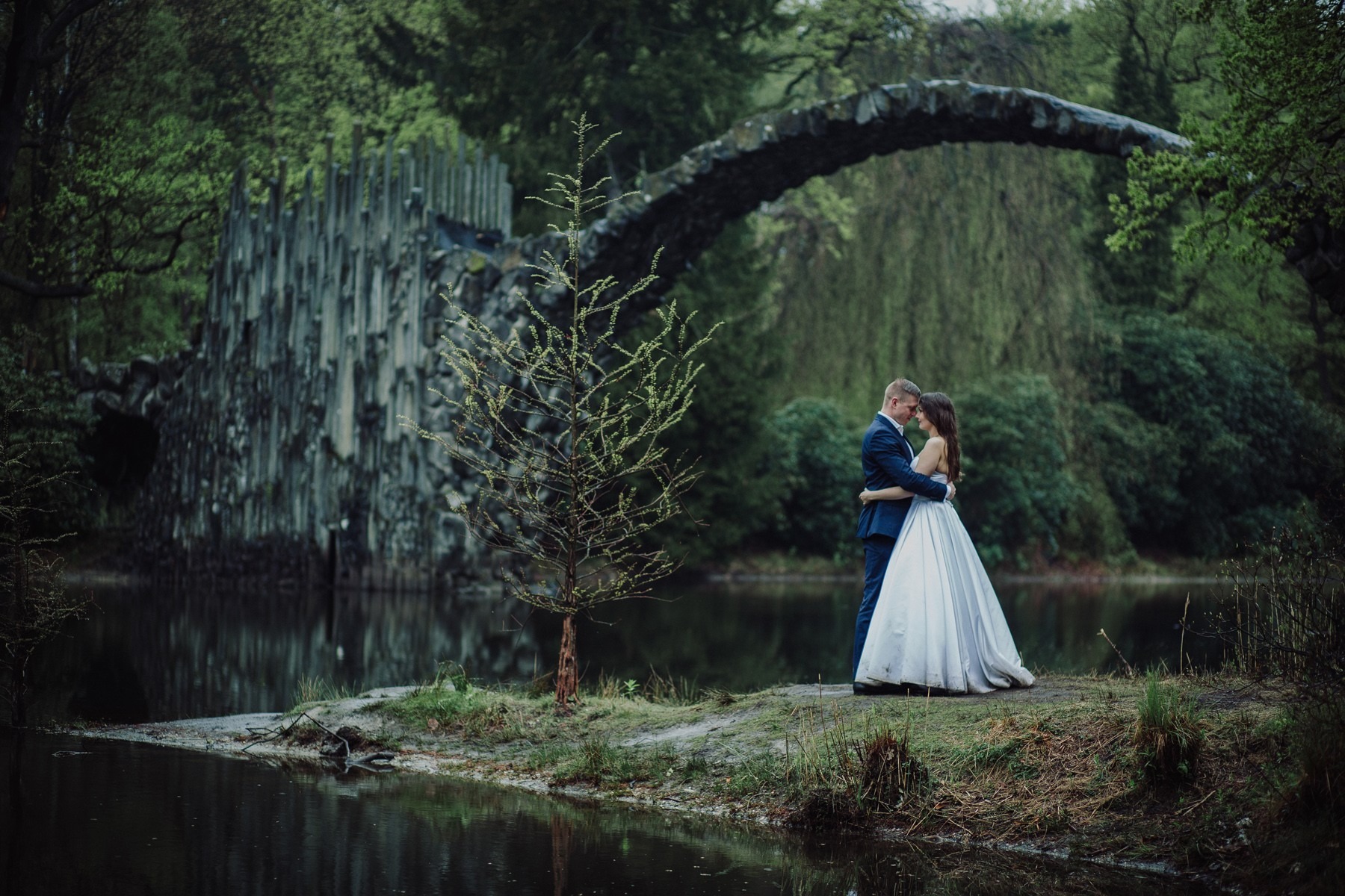 Para młoda obejmuje się na tle malowniczego mostu Devil's Bridge w Kromlau, Niemcy, z unikalnymi bazaltowymi formacjami skalnymi i wodą.