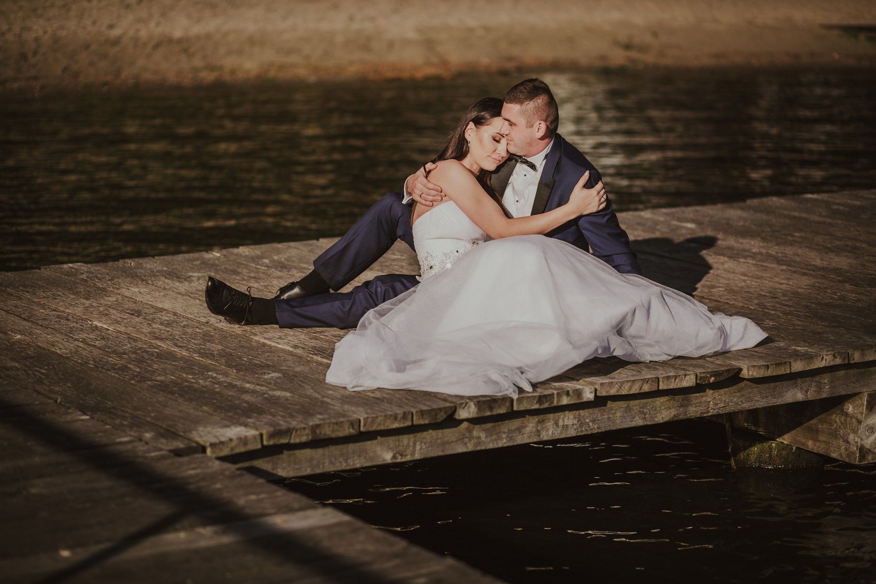 Para młoda siedzi na drewnianym pomoście nad wodą, w tle widoczne drzewa, panna młoda w białej sukni, pan młody w granatowym garniturze z muszką.