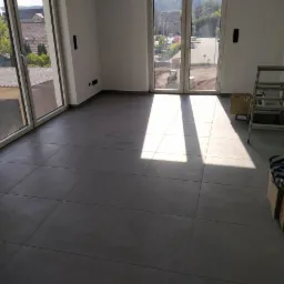 Wykończone wnętrze pokoju z szarymi, kwadratowymi płytkami podłogowymi, widoczne skrzynki z materiałami i drabina aluminiowa, wpadające promienie słoneczne.