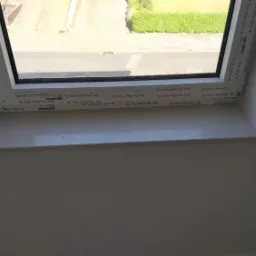 Biały parapet wewnętrzny z PCV pod oknem dachowym, widok na ulicę z zielenią w tle.