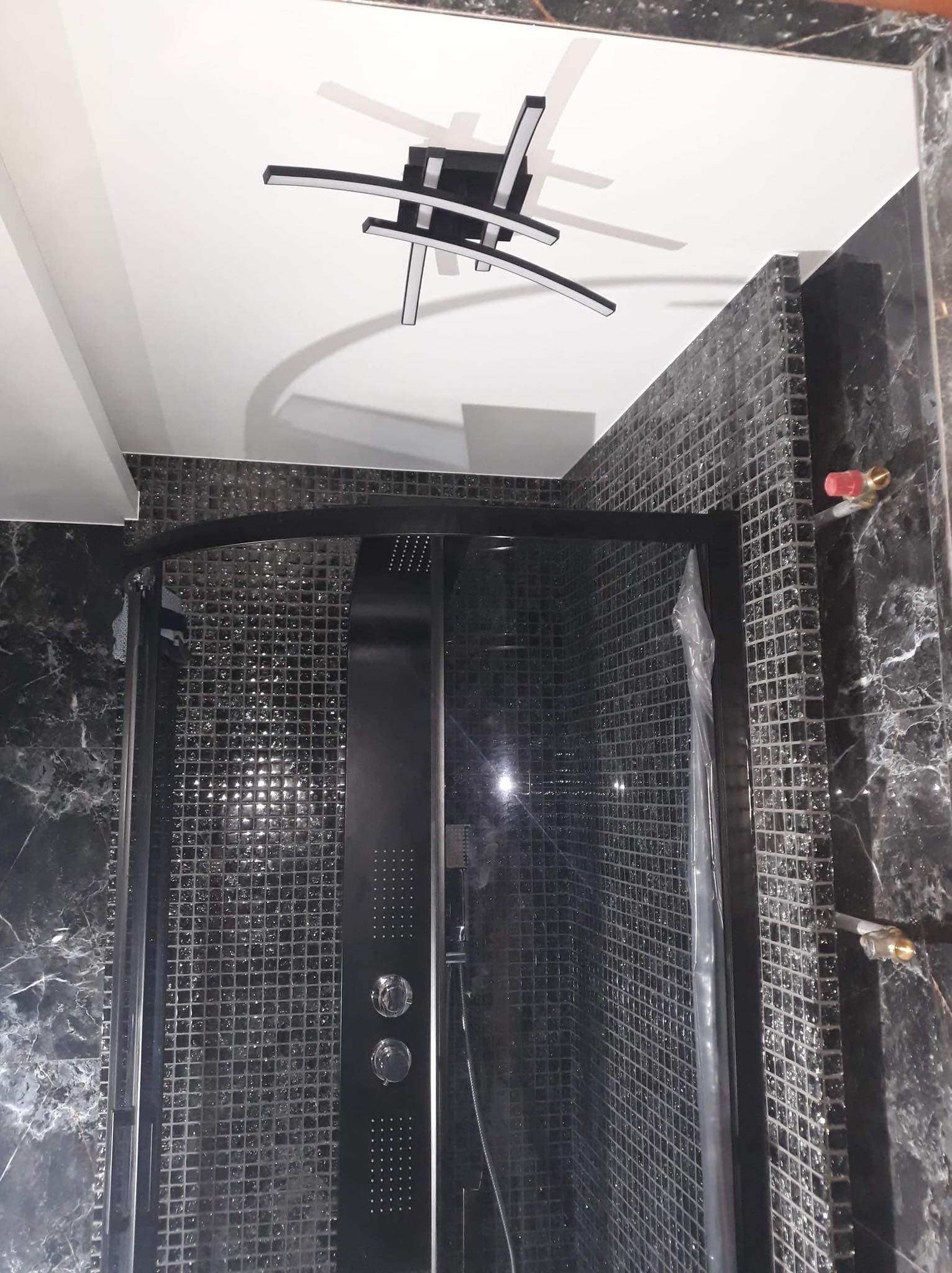 Kabina prysznicowa z czarnymi profilami i mozaiką w odcieniach szarości, oświetlona nowoczesną lampą sufitową.