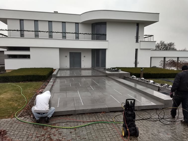 Układanie szarej, prostokątnej kostki brukowej na posesji nowoczesnego domu, widoczna myjka ciśnieniowa i dwaj pracownicy w trakcie pracy.