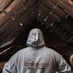 FRB Silesia - Osoba w bluzie z logo firmy budowlanej stojąca w ocieplonym wełną mineralną poddaszu z widocznymi elementami mocującymi izolację.
