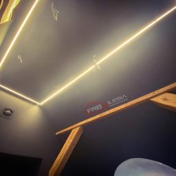 FRB Silesia - Nowoczesny sufit z podświetleniem LED w kształcie prostokąta, widoczne drewniane elementy konstrukcyjne poddasza i wentylator w rogu.