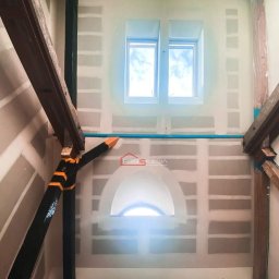 FRB Silesia - Poddasze w trakcie prac wykończeniowych: ściany z płyt kartonowo-gipsowych, drewniane belki konstrukcyjne pomalowane na czarno z pomarańczowymi akcentami, okno dachowe oraz łukowe okno poniżej...