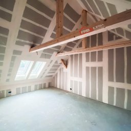 FRB Silesia - Poddasze w trakcie montażu płyt kartonowo-gipsowych, widoczne drewniane elementy konstrukcyjne i okno dachowe, ściany przygotowane pod dalsze prace wykończeniowe, logo firmy widoczne na belce.
