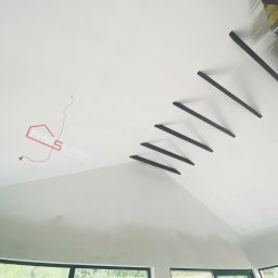 FRB Silesia - Białe, wyszpachlowane poddasze ze skośnym sufitem, dekoracyjne drewniane belki, logo firmy i wystające przewody elektryczne.