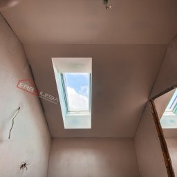 FRB Silesia - Poddasze w trakcie prac wykończeniowych, widoczne dwa okna dachowe z widokiem na błękitne niebo, ściany i sufit pokryte gładzią szpachlową przed malowaniem, wystające przewody elektryczne, logo...
