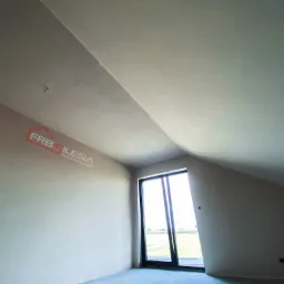 Poddasze po gładziowaniu, widoczne ściany pomalowane na jasny kolor, okno balkonowe i fragment logo firmy na ścianie.