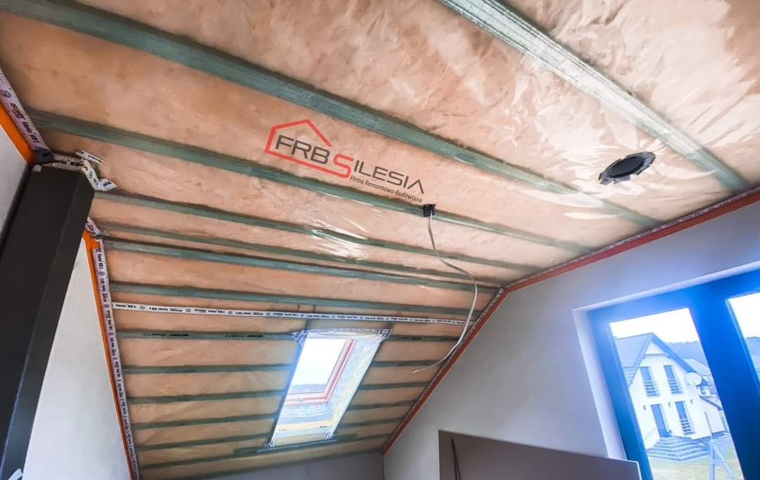 Poddasze z ociepleniem wełną mineralną, zabezpieczone folią, widoczne drewniane belki konstrukcyjne oraz okno dachowe, logo firmy FRB Silesia na folii.