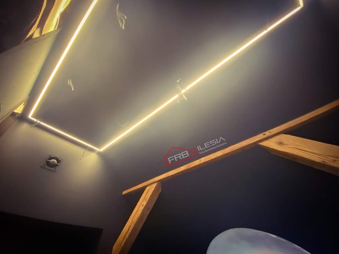 Nowoczesny sufit z podświetleniem LED w kształcie prostokąta, widoczne drewniane elementy konstrukcyjne poddasza i wentylator w rogu.