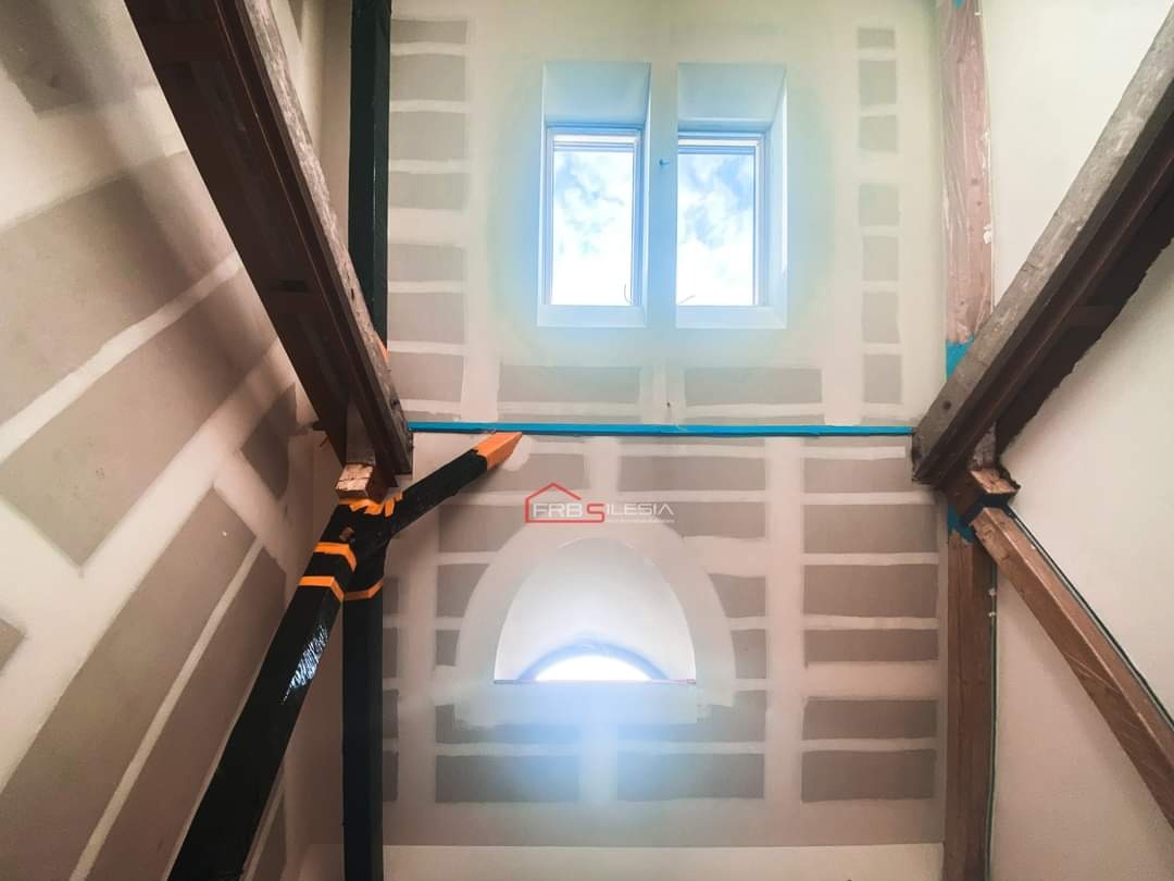 Poddasze w trakcie prac wykończeniowych: ściany z płyt kartonowo-gipsowych, drewniane belki konstrukcyjne pomalowane na czarno z pomarańczowymi akcentami, okno dachowe oraz łukowe okno poniżej...