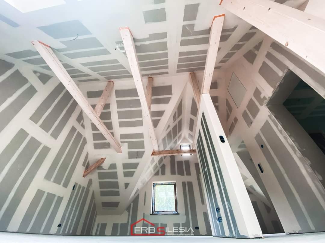 Poddasze z widoczną konstrukcją drewnianą i płytami kartonowo-gipsowymi przed szpachlowaniem, małe okno w ścianie szczytowej, widoczny fragment logo firmy w rogu