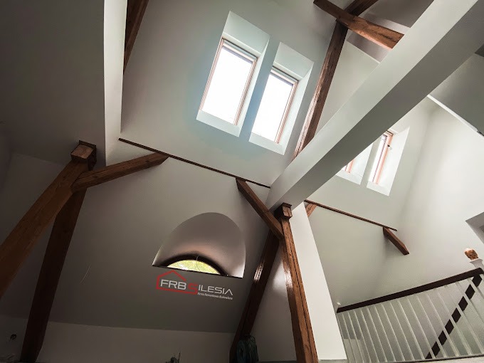 Wnętrze poddasza po remoncie z widocznymi drewnianymi belkami konstrukcyjnymi, oknami dachowymi i wykończonymi ścianami w kolorze białym. Widoczny fragment schodów z białą balustradą. W dolnej...