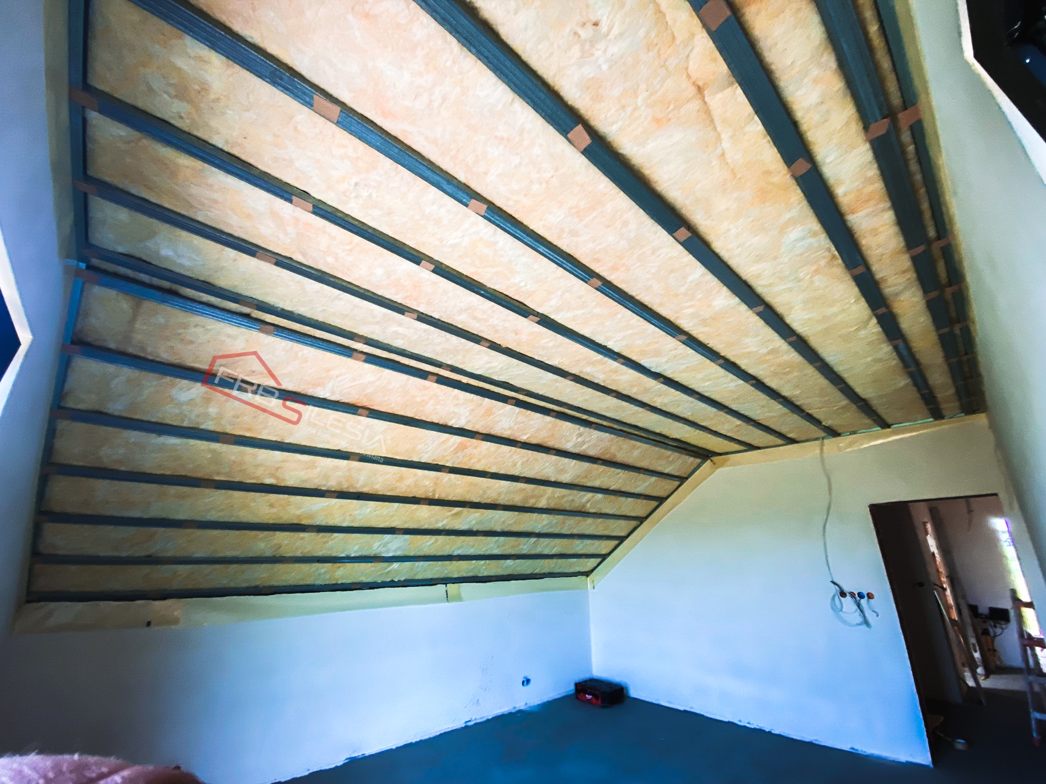 Skośny sufit poddasza w trakcie ocieplania wełną mineralną, widoczne metalowe profile konstrukcyjne i częściowo wykończona ściana, z wystającymi przewodami elektrycznymi.