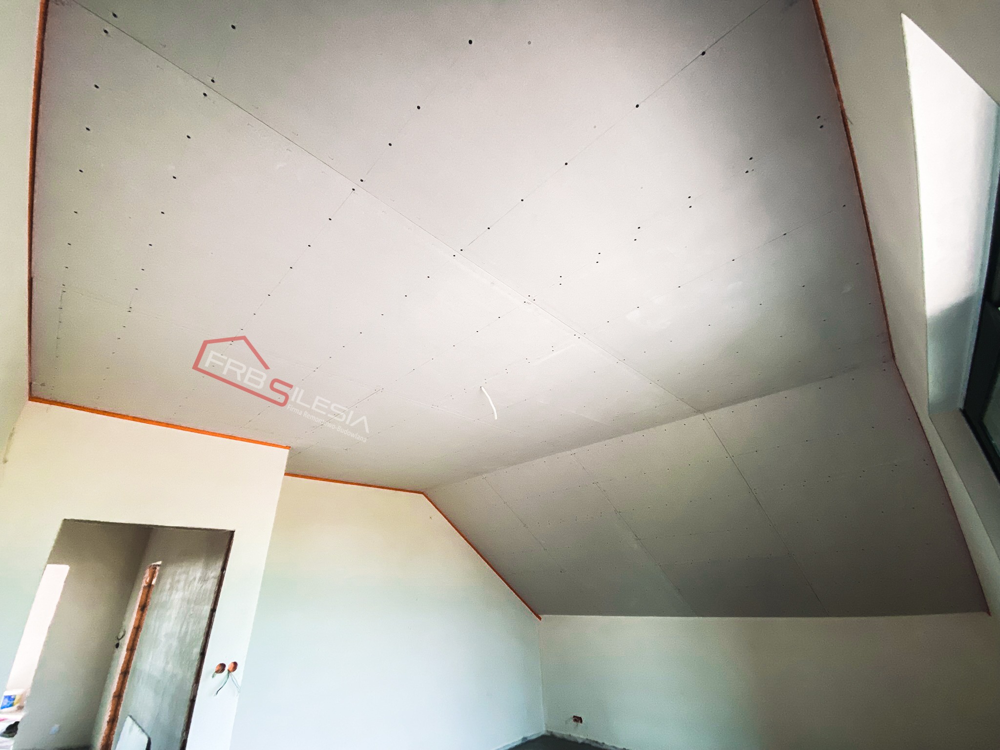 Poddasze z zamontowanymi płytami karton-gips, widoczne wkręty, taśma wzmacniająca na łączeniach i fragment logo firmy na suficie, widok z dołu.