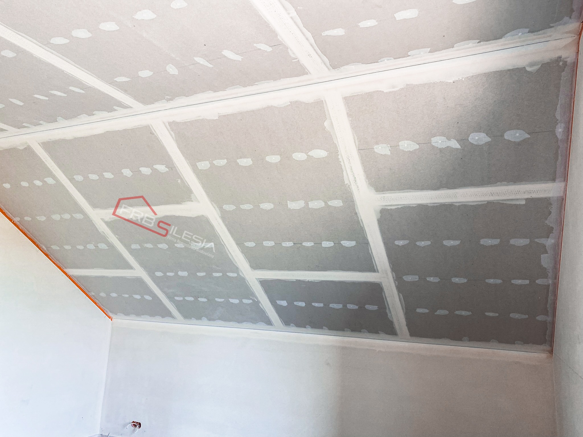 Skośny sufit z płyt karton gips, widoczne łączenia płyt i ślady po szpachlowaniu, logo firmy na suficie.