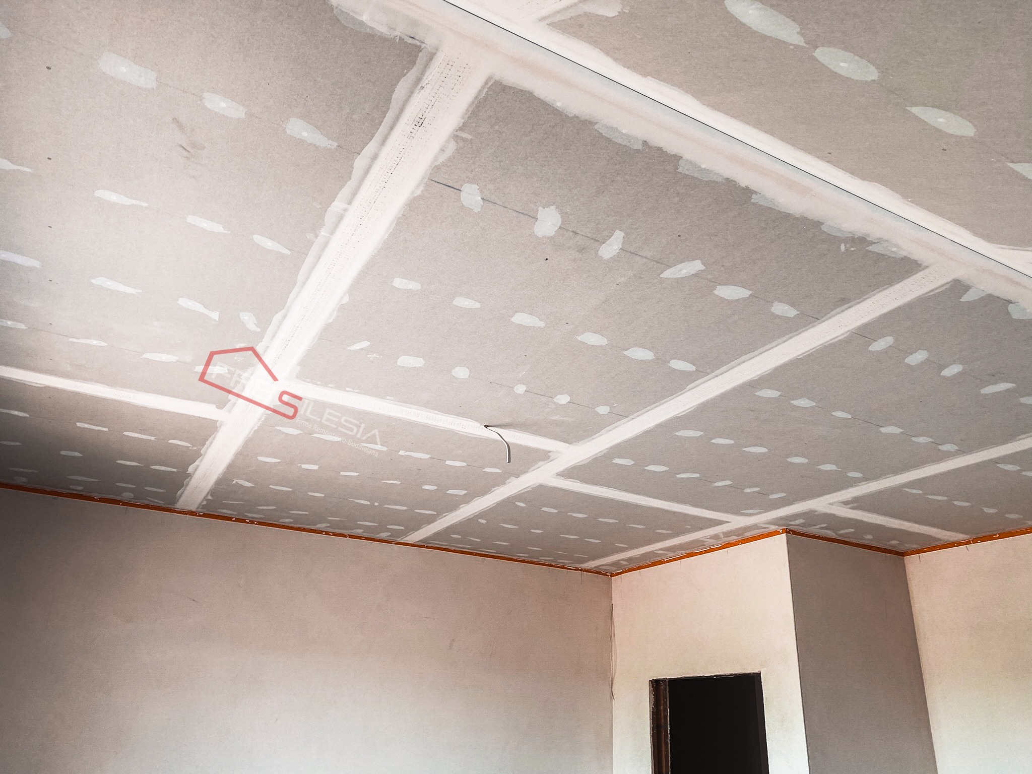 Sufit z płyt kartonowo-gipsowych w trakcie montażu, widoczne łączenia płyt zaszpachlowane, przewody elektryczne wystające z sufitu, logo firmy w rogu.