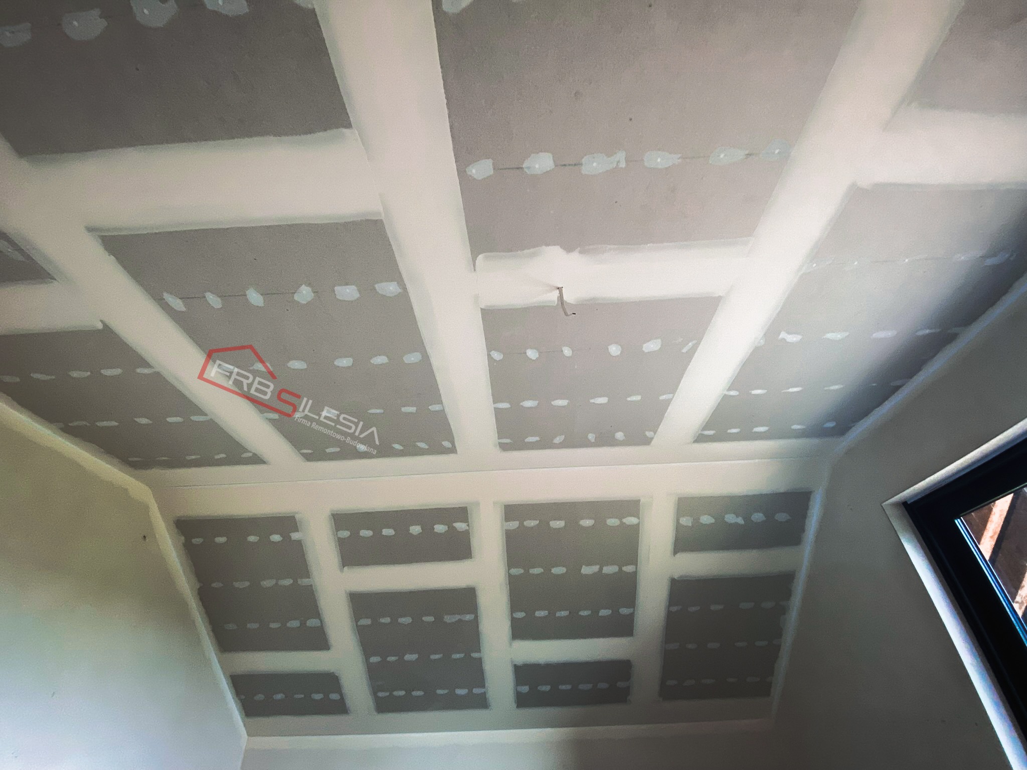 Sufit podwieszany z płyt kartonowo-gipsowych w trakcie montażu, widoczne profile i łączenia, szpachlowane miejsca łączeń, logo firmy w lewym górnym rogu.