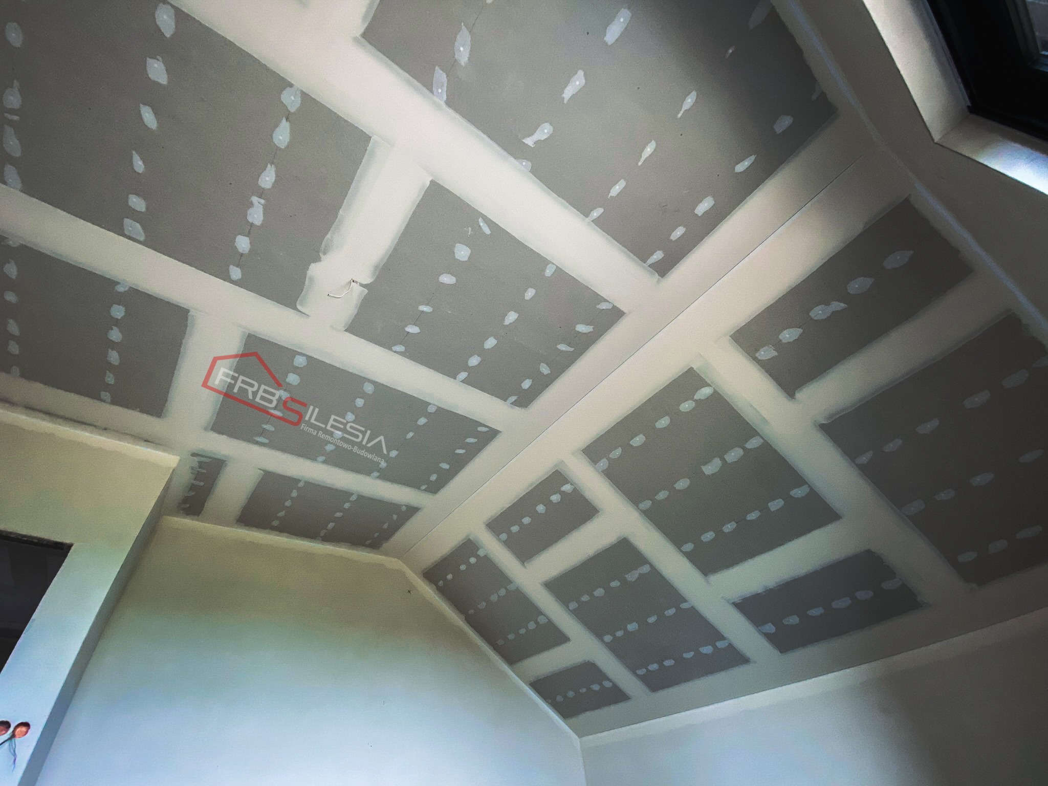 Skośny sufit poddasza z zamontowanymi płytami kartonowo-gipsowymi, widoczne łączenia płyt wypełnione masą szpachlową, logo firmy w rogu sufitu.