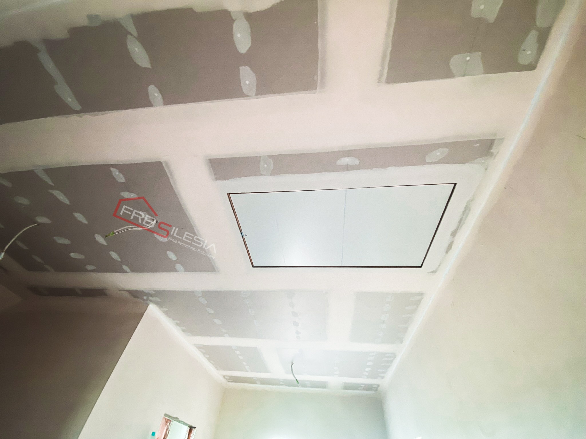 Montaż sufitu z płyt kartonowo-gipsowych z otworem rewizyjnym w Rudzie Śląskiej, widoczne łączenia płyt przed szpachlowaniem, logo firmy w rogu.