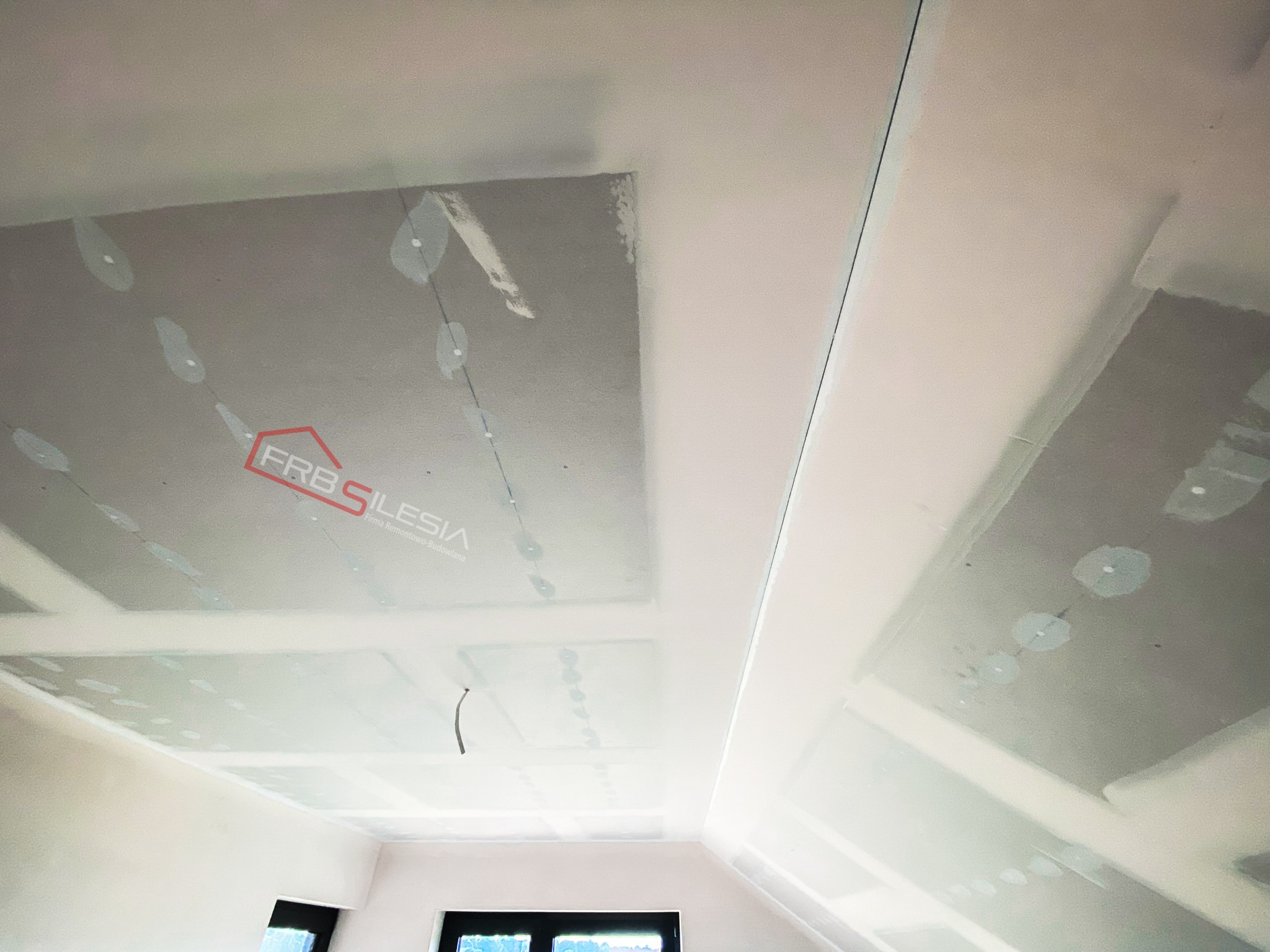 Montaż płyt kartonowo-gipsowych na suficie podwieszanym, widoczne łączenia i logo firmy FRB Silesia.