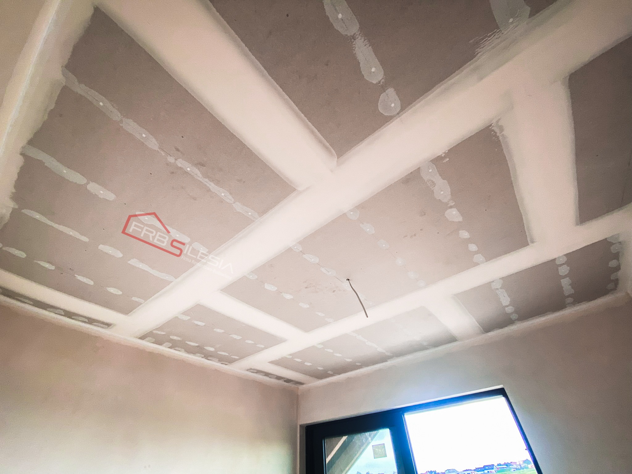 Sufit z płyt kartonowo-gipsowych z widocznymi łączeniami wypełnionymi masą szpachlową, widok z dołu, częściowo widoczna ściana i okno.