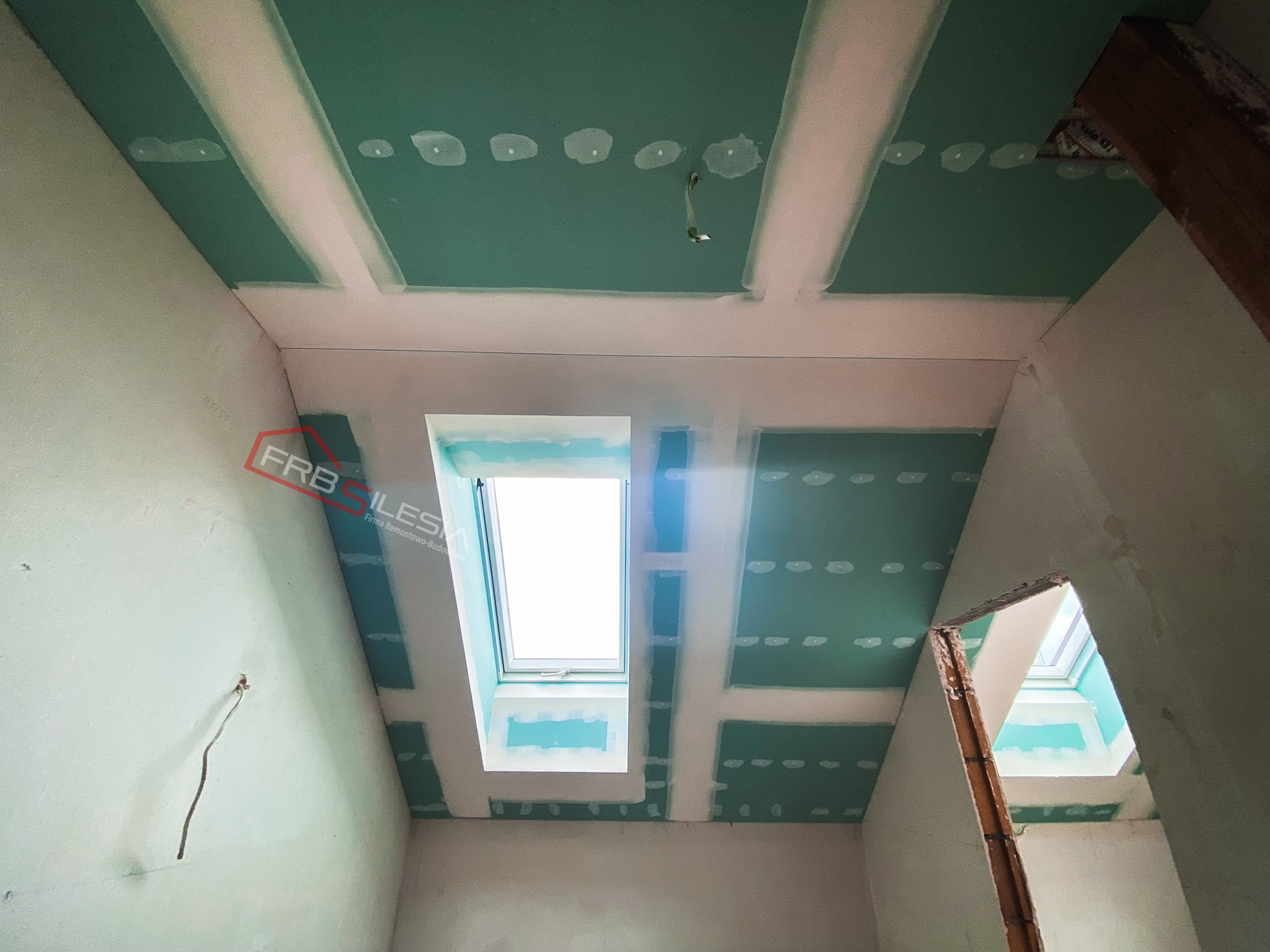 Poddasze z oknem dachowym w trakcie montażu płyt karton gips, widoczne szpachlowane łączenia i śruby, z fragmentem instalacji elektrycznej wystającej ze ściany.