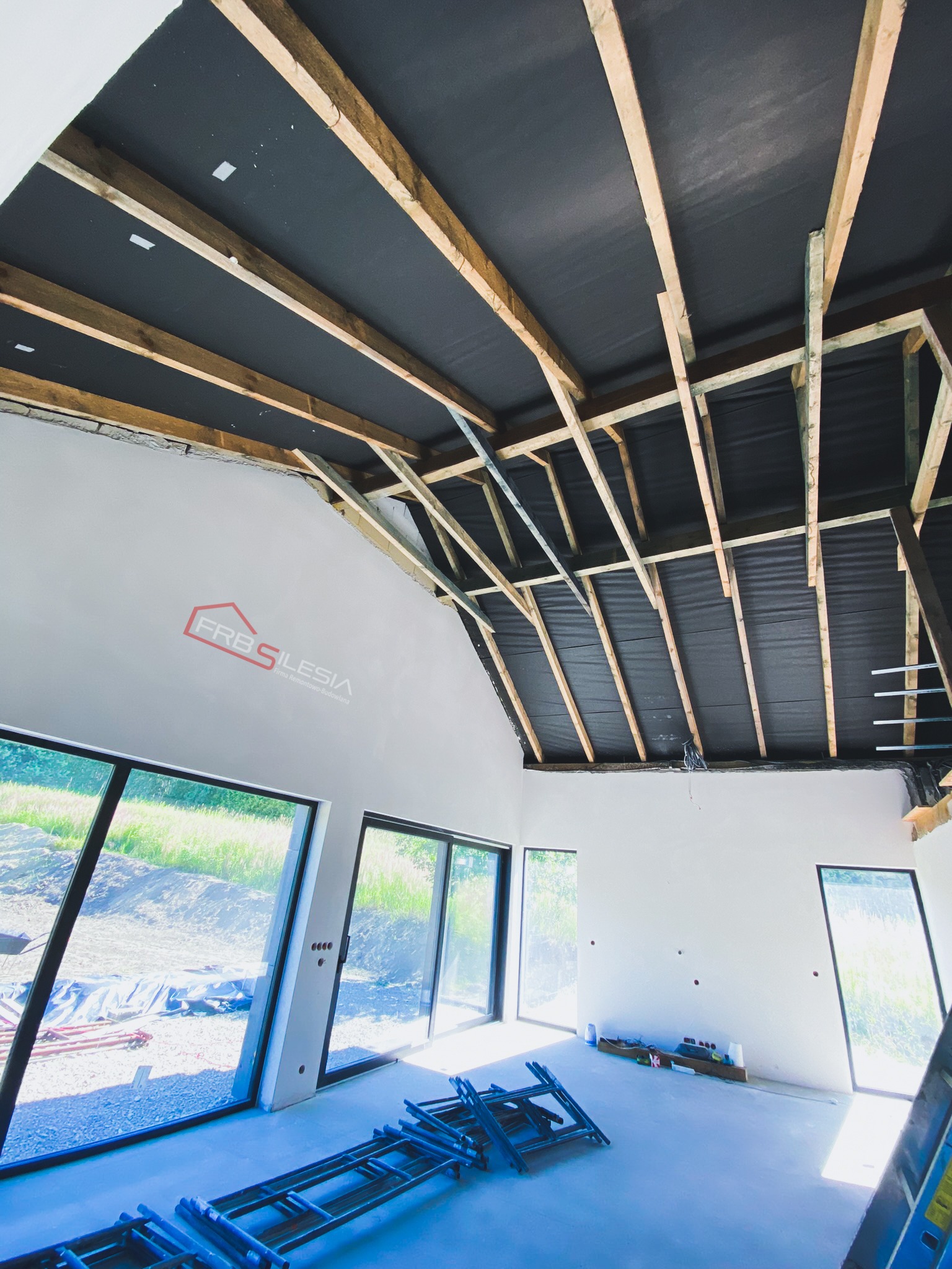 Wnętrze w stanie surowym: widoczne drewniane belki stropowe z czarną membraną, białe ściany z logo firmy FRB Silesia, duże okna z widokiem na zewnątrz oraz rozłożone rusztowanie na podłodze.