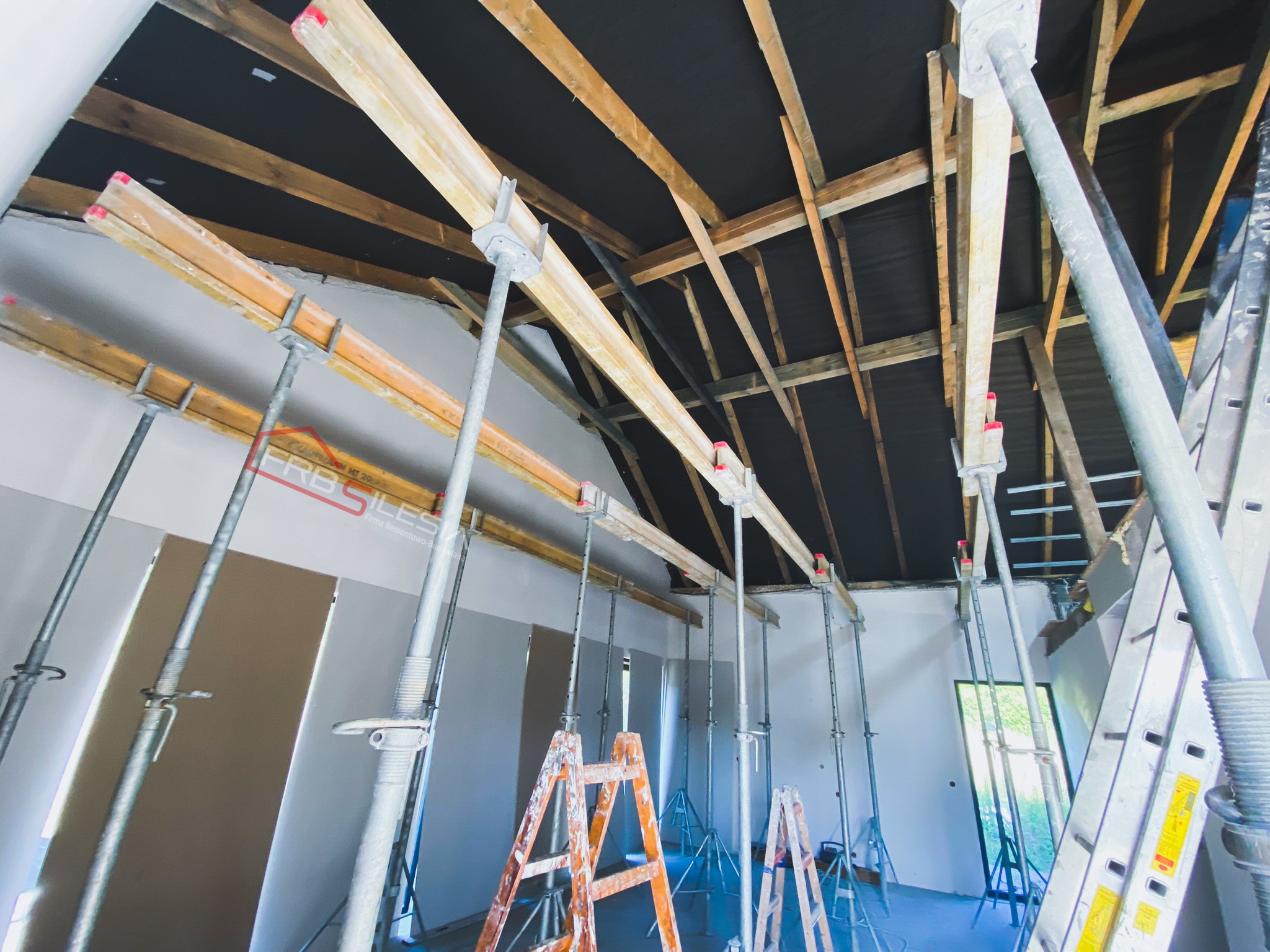Wnętrze w trakcie montażu płyt kartonowo-gipsowych na poddaszu, widoczne stelaże, podpory teleskopowe, drabiny i czarna membrana dachowa.