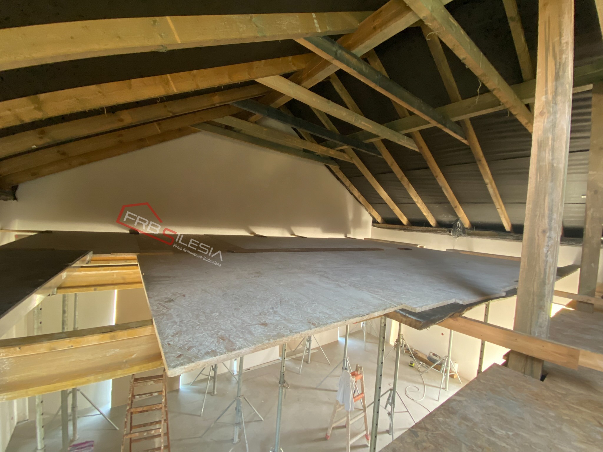 Poddasze w trakcie adaptacji: widoczne drewniane belki konstrukcyjne, czarna folia, płyty OSB tworzące podłogę, metalowe podpory i drabina. W tle logo firmy budowlanej.