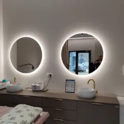 Dwa okrągłe lustra z podświetleniem LED nad umywalkami w gabinecie kosmetycznym, widoczne wyposażenie i odbicie osoby w jednym z luster.