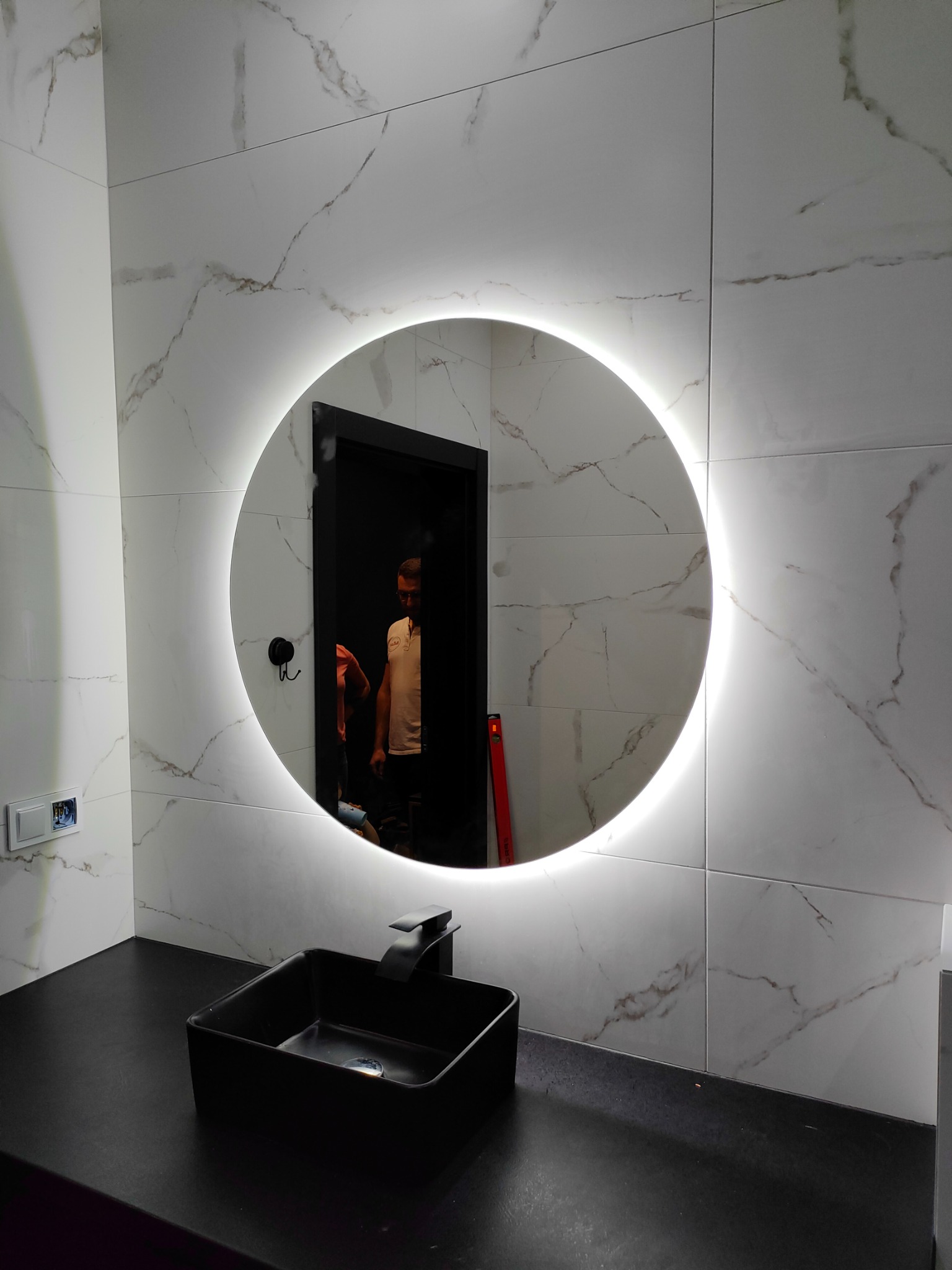 Okrągłe lustro podświetlane LED w nowoczesnej łazience z marmurowymi płytkami, czarną umywalką nablatową i widocznym odbiciem osoby.