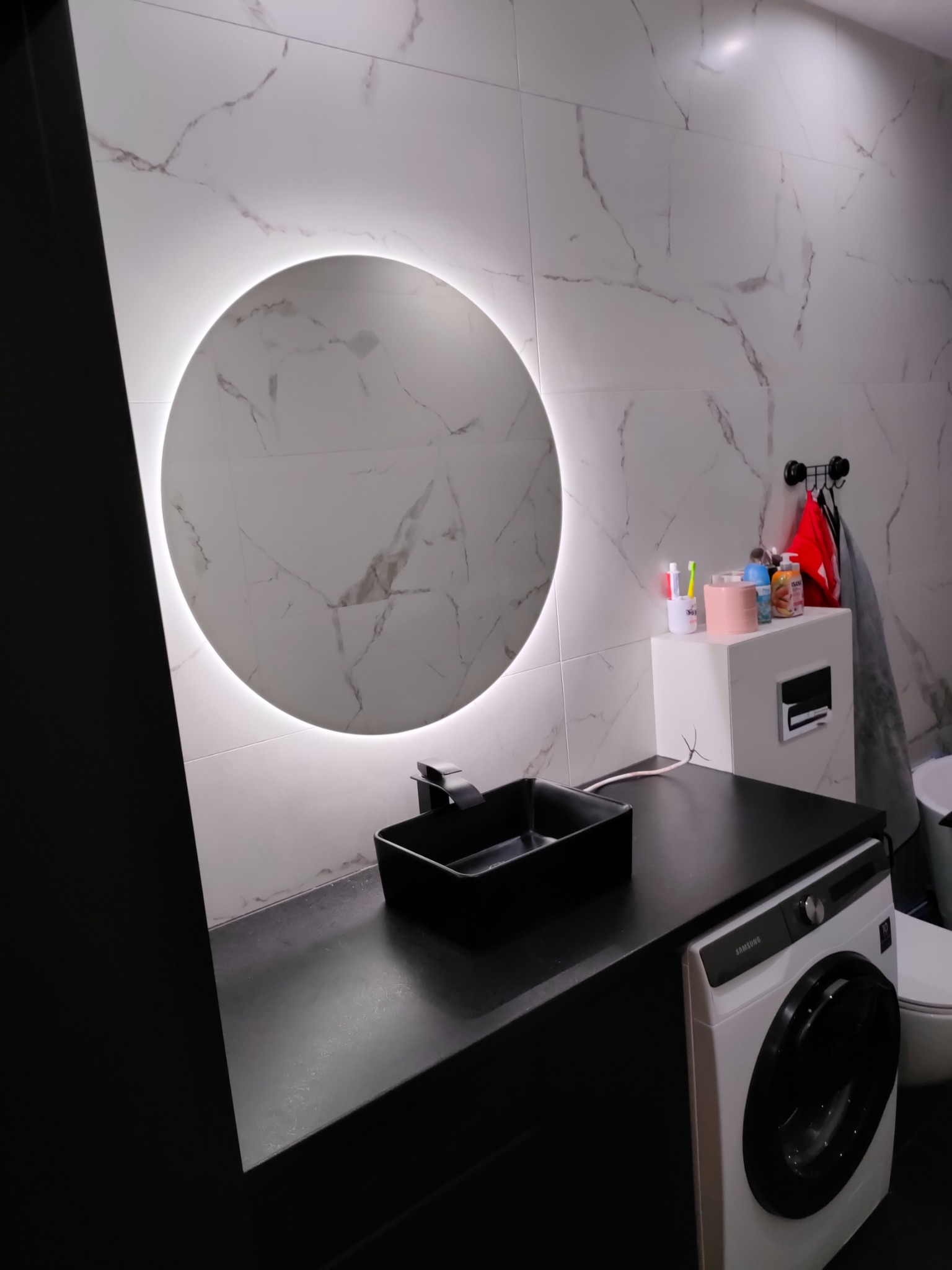 Nowoczesna łazienka z okrągłym lustrem podświetlanym LED, czarną umywalką nablatową i blatem oraz pralką Samsung w zabudowie.