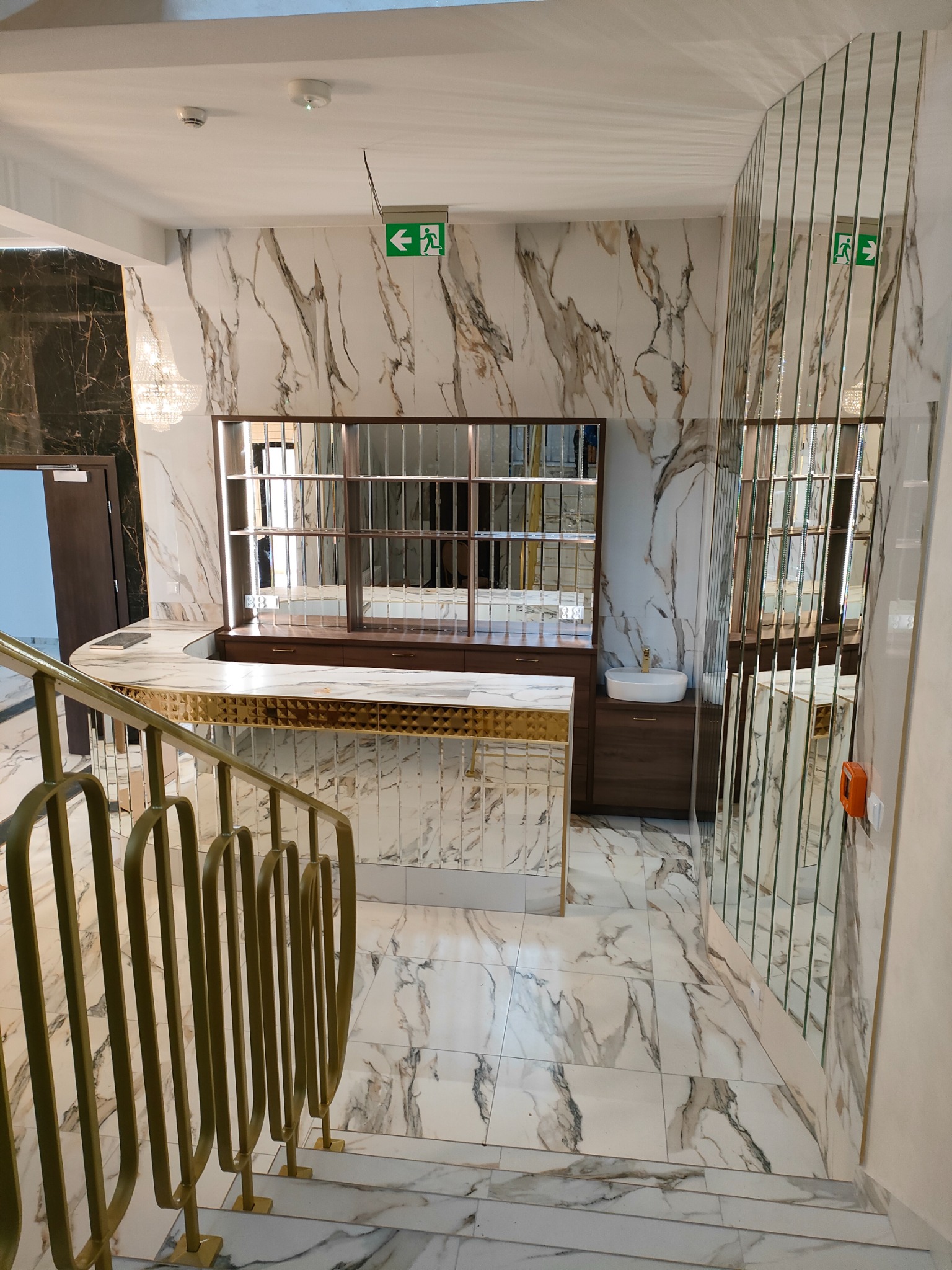 Nowoczesne wnętrze z marmurowymi płytkami i złotą balustradą schodową, widoczne lustrzane panele i recepcja z umywalką.