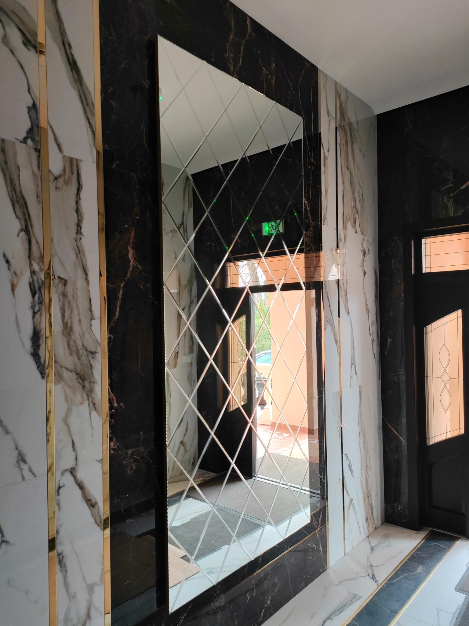 Dekoracyjne lustro ścienne z romboidalnych elementów, otoczone marmurowymi płytami z złotymi akcentami, widoczne drzwi w tle.