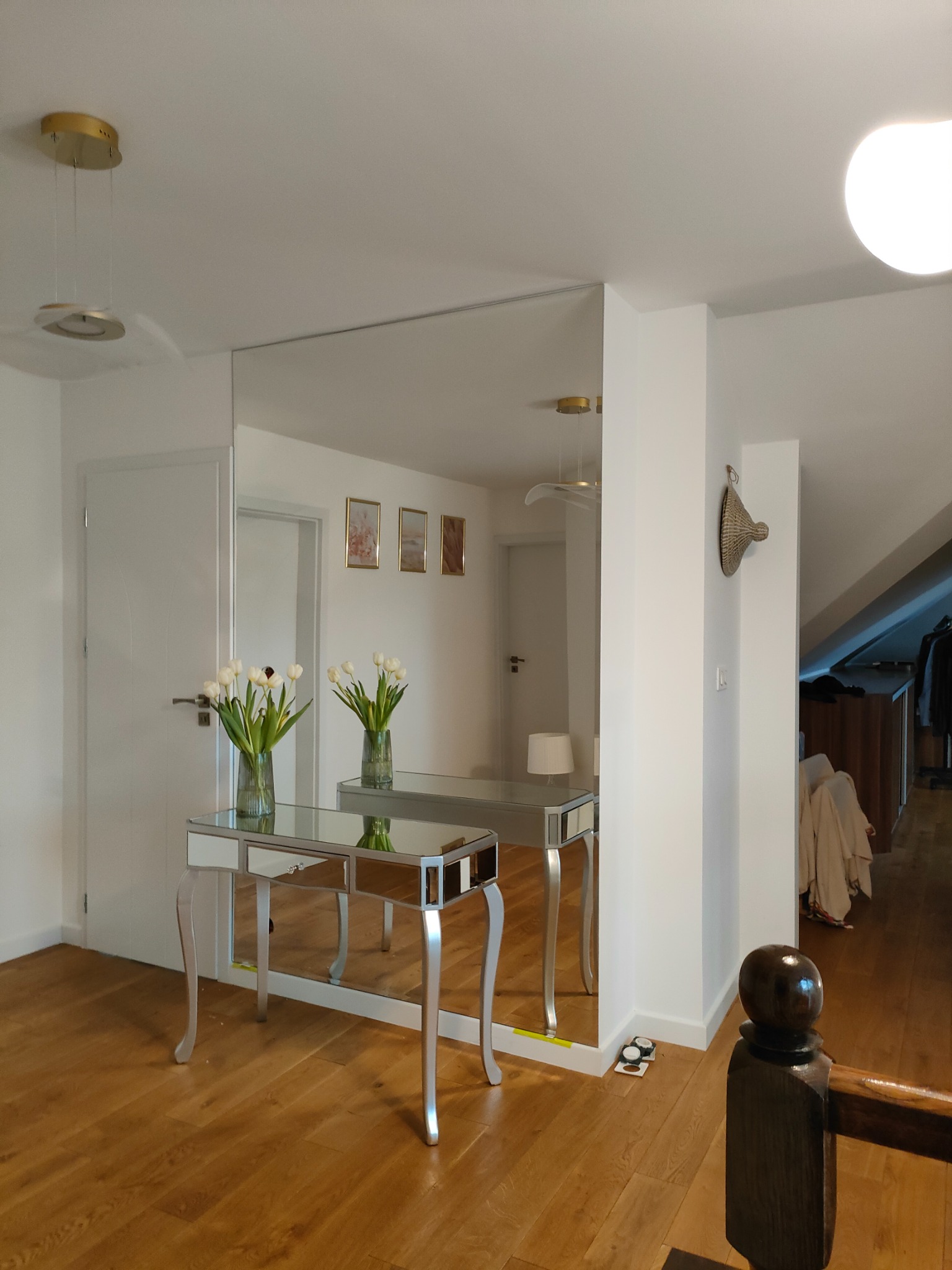 Duże lustro na ścianie w przedpokoju, odbijające stolik z lustrzanymi elementami i wazonami z białymi tulipanami. Widoczne drewniane podłogi i schody.
