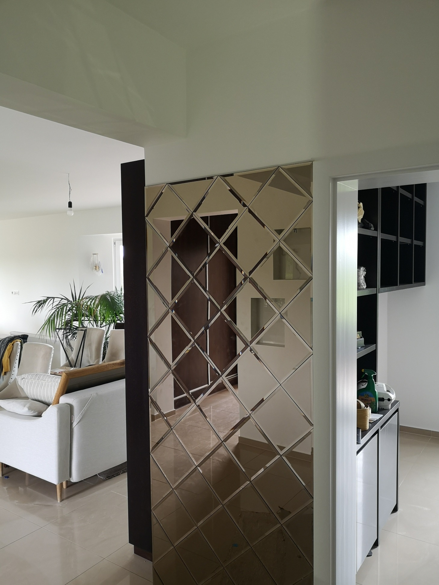 Dekoracyjna ściana z lustrzanych płytek w kształcie rombów, odbijająca wnętrze salonu z sofą i roślinami, oddzielona od pomieszczenia z szafką.